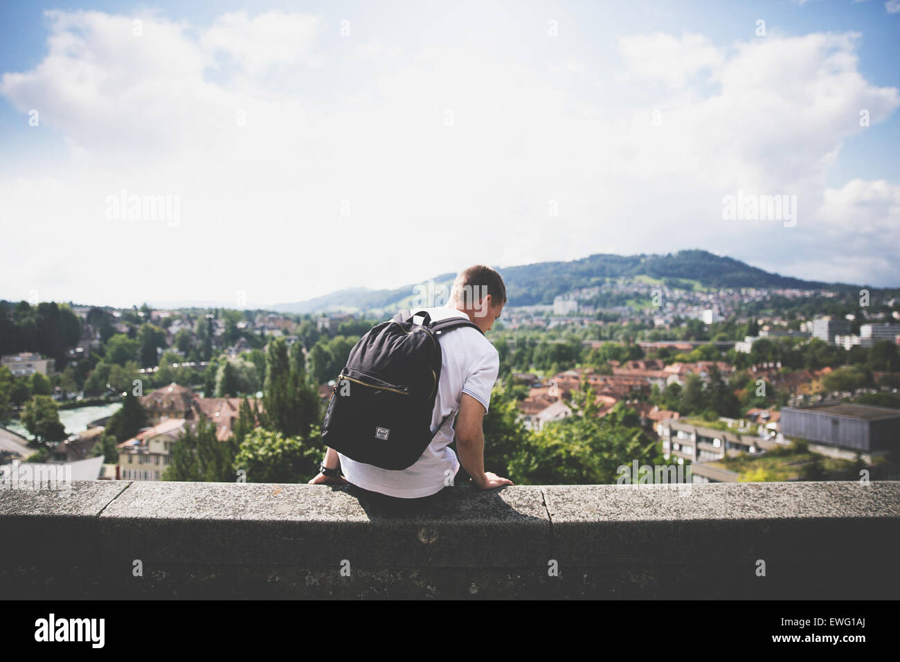 Un uomo con uno zaino siede su una sporgenza, affacciato sulla città sottostante. Questa immagine trasmette un senso di solitudine e contemplazione, mentre la persona prende in considerazione la vista espansiva del paesaggio urbano. Il contrasto tra l'individuo e la città evidenzia l'equilibrio tra riflessione personale e il trambusto della vita cittadina. Foto Stock