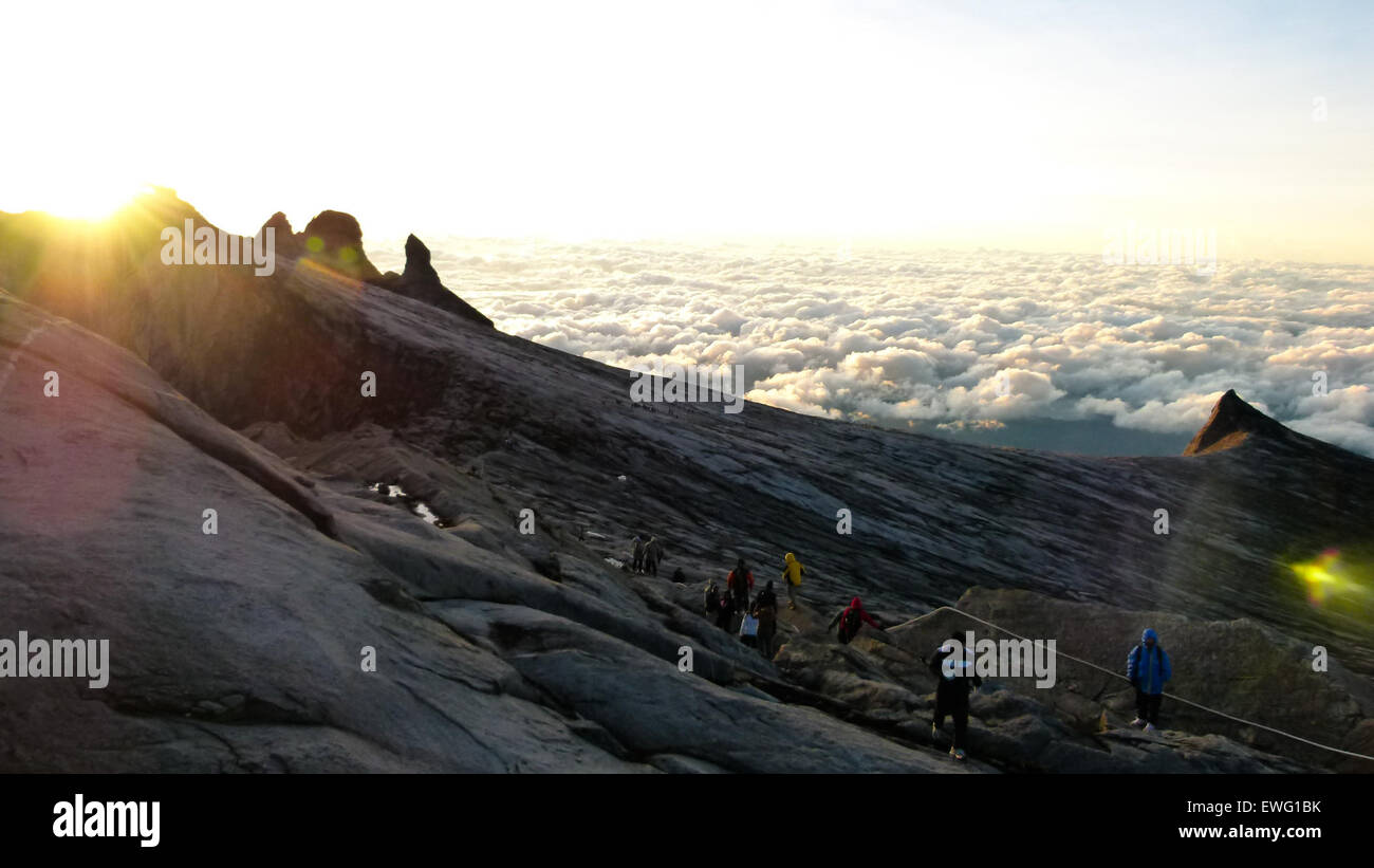Gli scalatori salgono una vetta sopra le nuvole, mostrando la vista mozzafiato del paesaggio circostante. Gli scalatori navigano sul terreno roccioso mentre sono circondati dalla luce solare e da un cielo esteso. La scena enfatizza le sfide e i premi dell'arrampicata in montagna, mettendo in evidenza sia lo sforzo fisico che la bellezza naturale degli ambienti ad alta quota. Foto Stock
