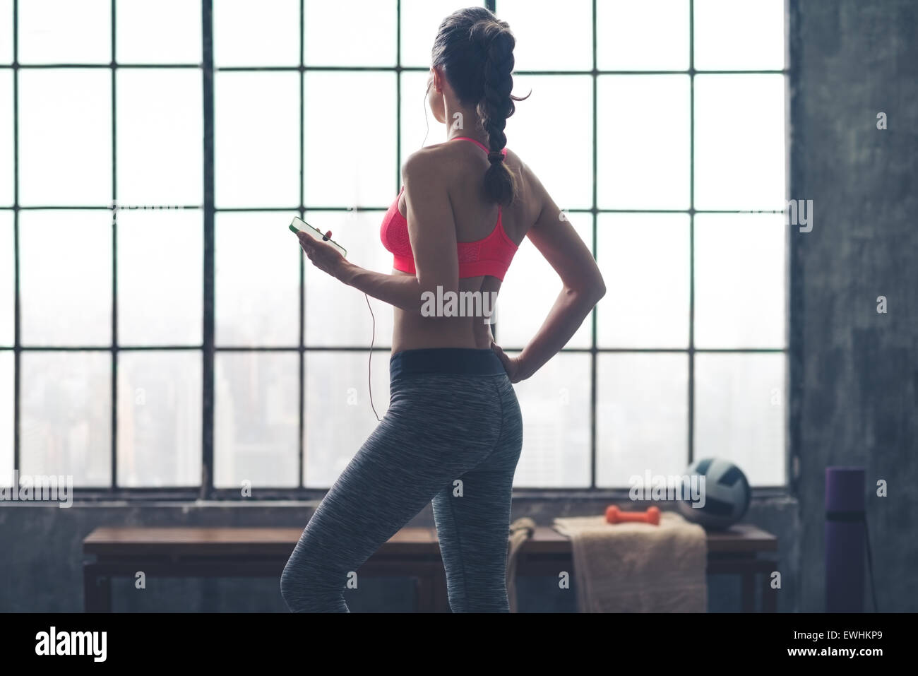 Una donna sta con una mano sulla sua anca, rilassante e guardando fuori della finestra, tenendo il suo dispositivo e ascolto di musica. Foto Stock