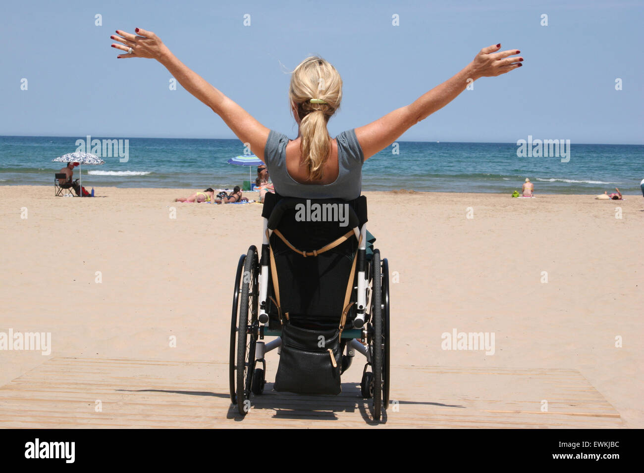 Una donna su una sedia a rotelle con le braccia aperte su una spiaggia guardando l'oceano Foto Stock