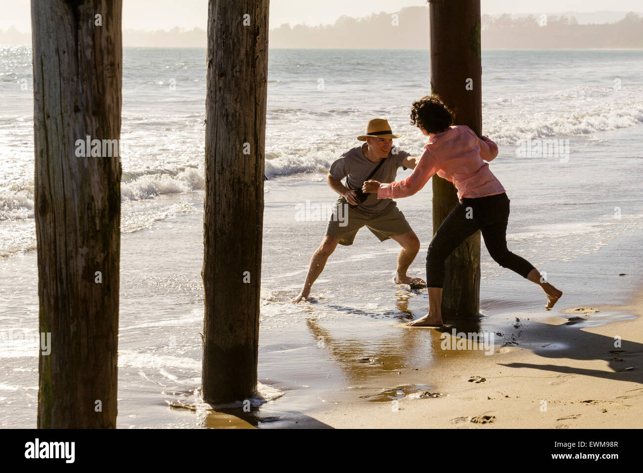 Uomo di mezza età e la donna giocosi e si rincorrono intorno al Boardwalk piloni in spiaggia Foto Stock