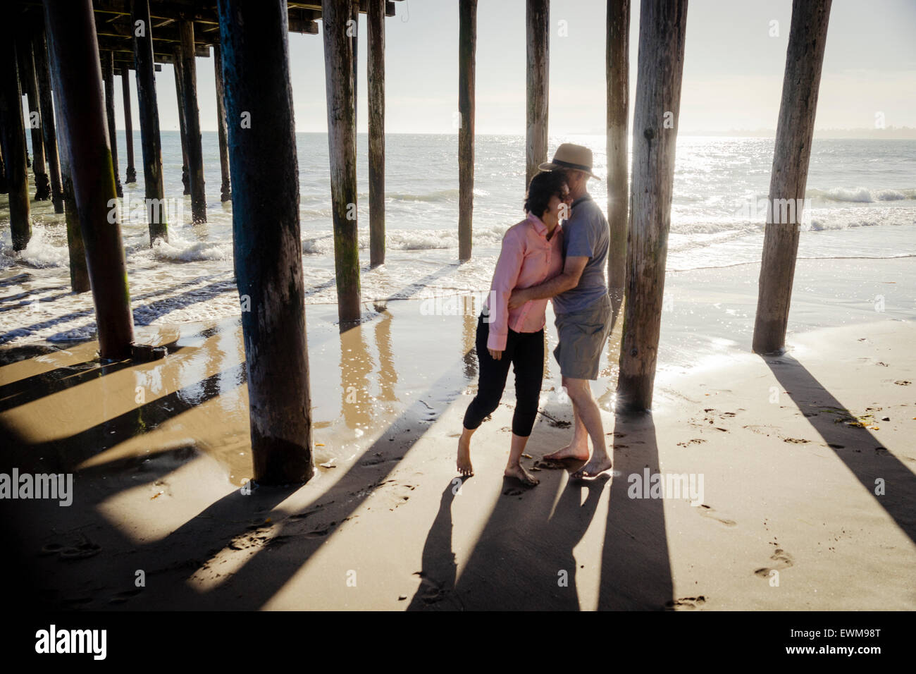 Uomo di mezza età e la donna abbracciando casualmente dopo che si rincorrono sotto il Boardwalk piloni in spiaggia Foto Stock