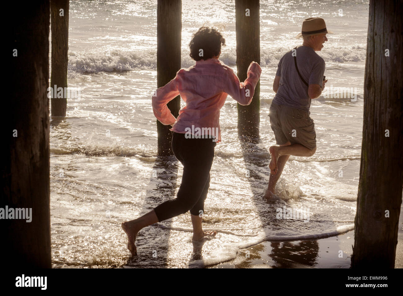 La donna a caccia di un uomo in spiaggia, schizzi attraverso acqua sotto il Boardwalk piloni Foto Stock
