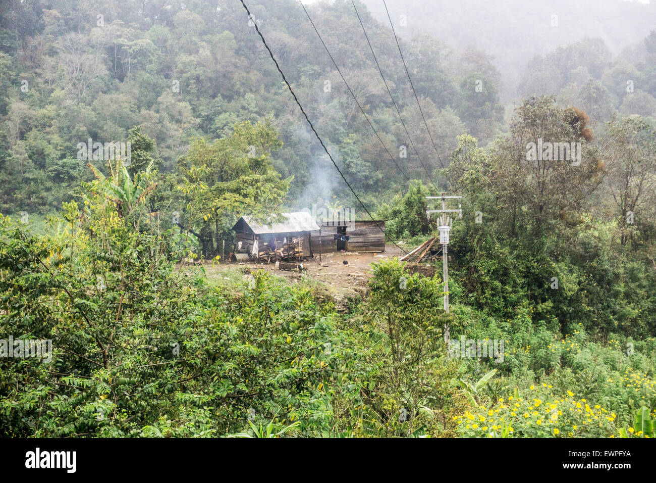 2 Stagno grezzo con tetto di baracche di legno accanto ad alta tensione linea e fumo di drifting che testimoniano la vita di sussistenza rurali in stato di Chiapas Foto Stock