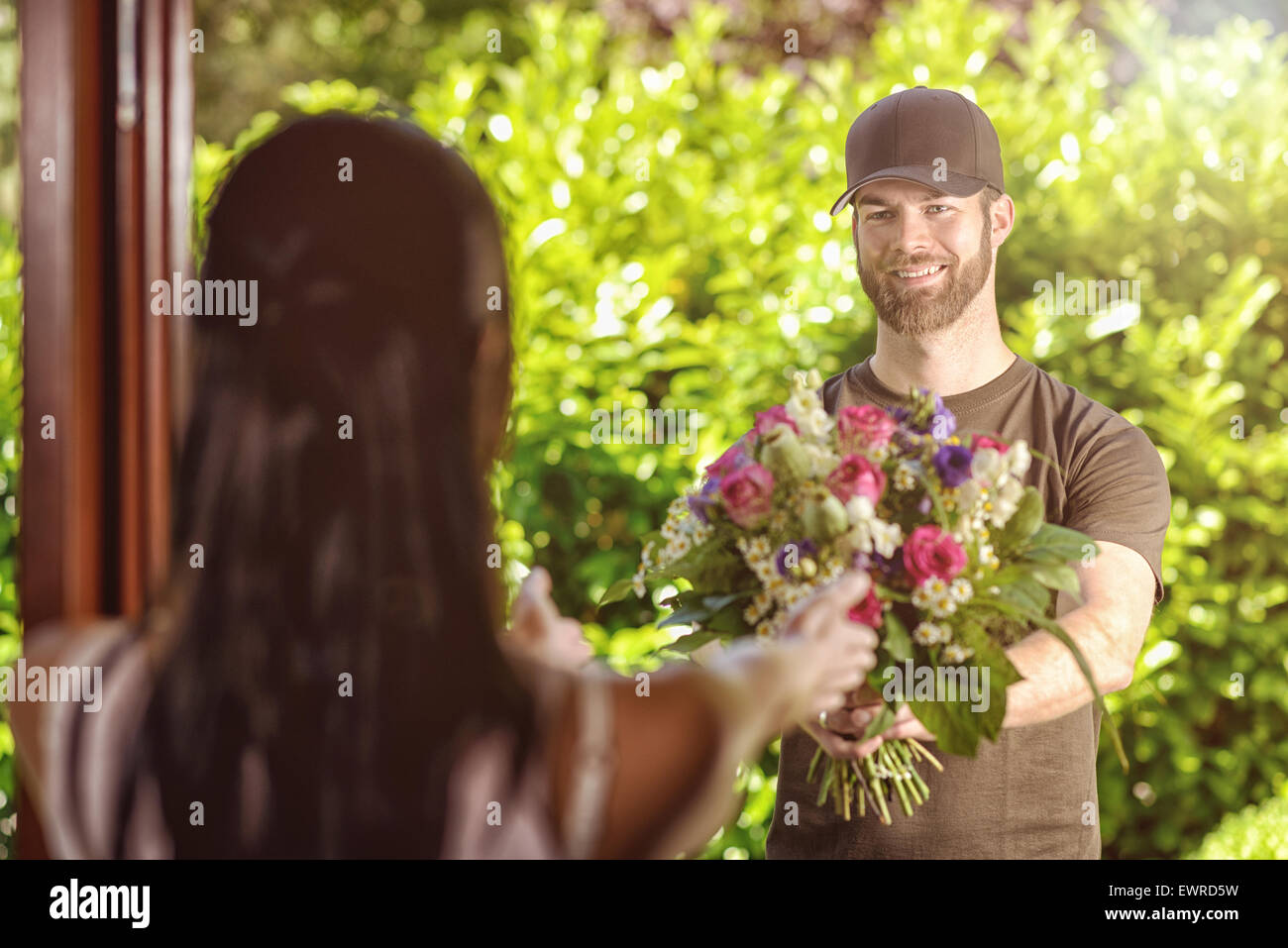 Sorridente barbuto 20s uomo che indossa tappo marrone e marrone t-shirt consegna fiori alla porta di giovani brunette femmina. Oltre la spalla Foto Stock