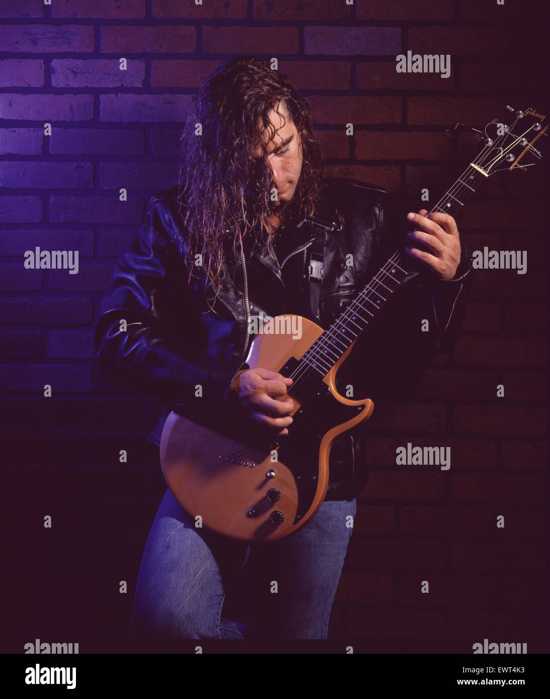 Chitarrista Rock con lunghi capelli biondi, indossa una giacca di pelle, suonare la chitarra elettrica contro un muro di mattoni. Foto Stock