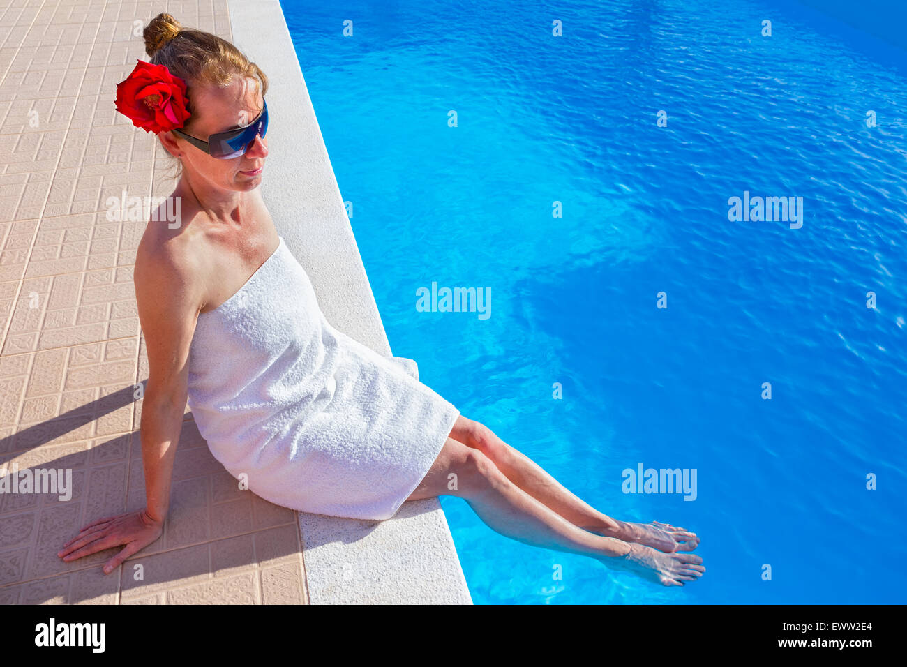 Donna europea che indossa asciugamano bianco rosa rossa e occhiali da sole tenendo le gambe in blu piscina Foto Stock