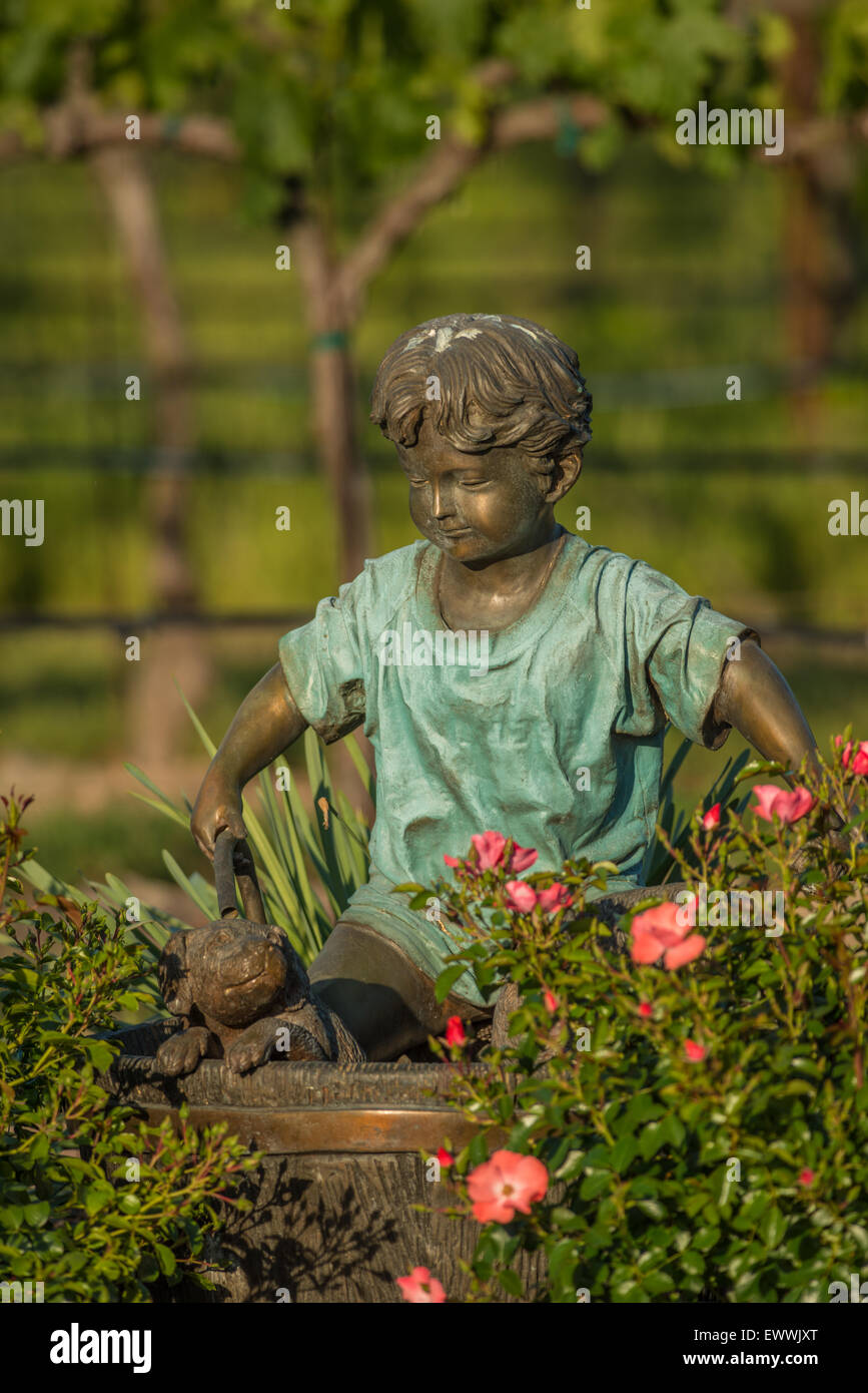 Ragazzo di bronzo e cane giardino statua collocata nel giardino fiorito vicino vigneto Foto Stock