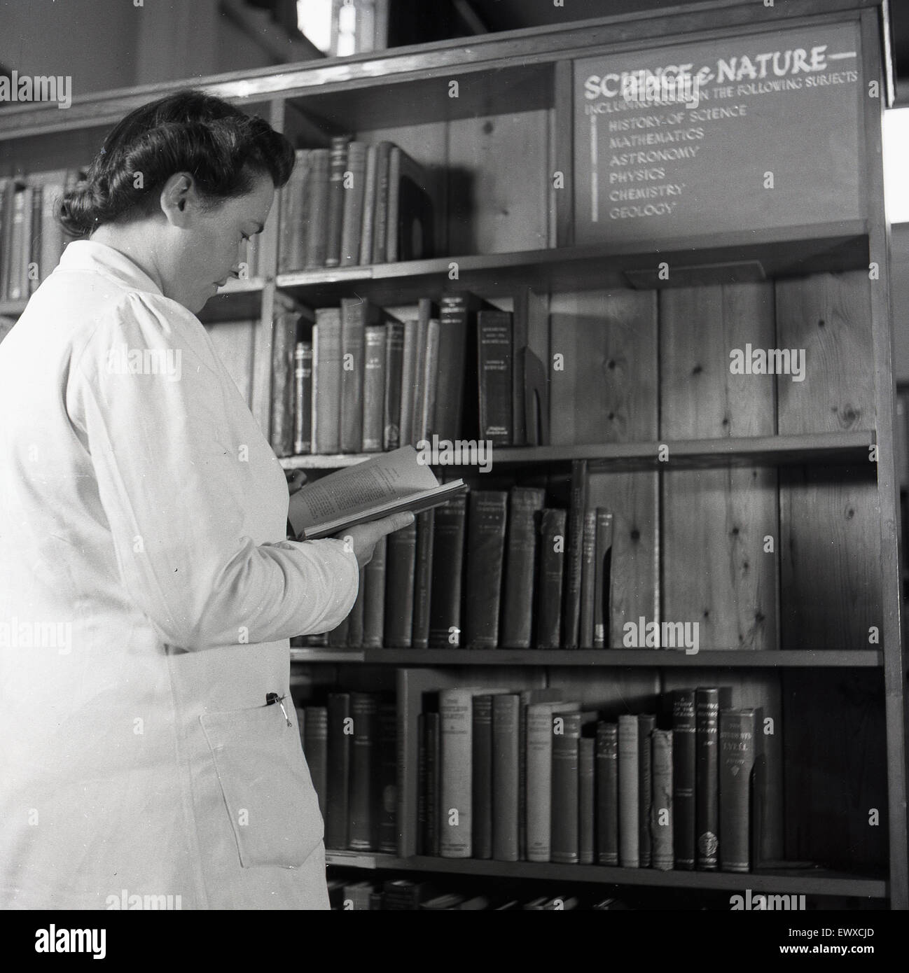 Anni '1950, storica, una signora con un cappotto bianco, una penna in tasca laterale, in una biblioteca pubblica, che guarda un libro nella sezione Scienza e natura. Foto Stock
