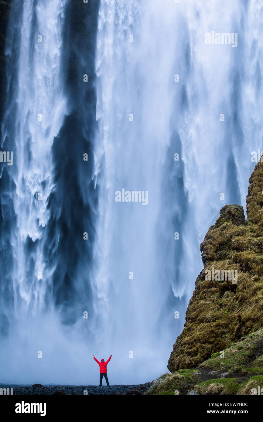 Uomo con le braccia in aria stando ai piedi della Cascata Skogafoss, Islanda Foto Stock