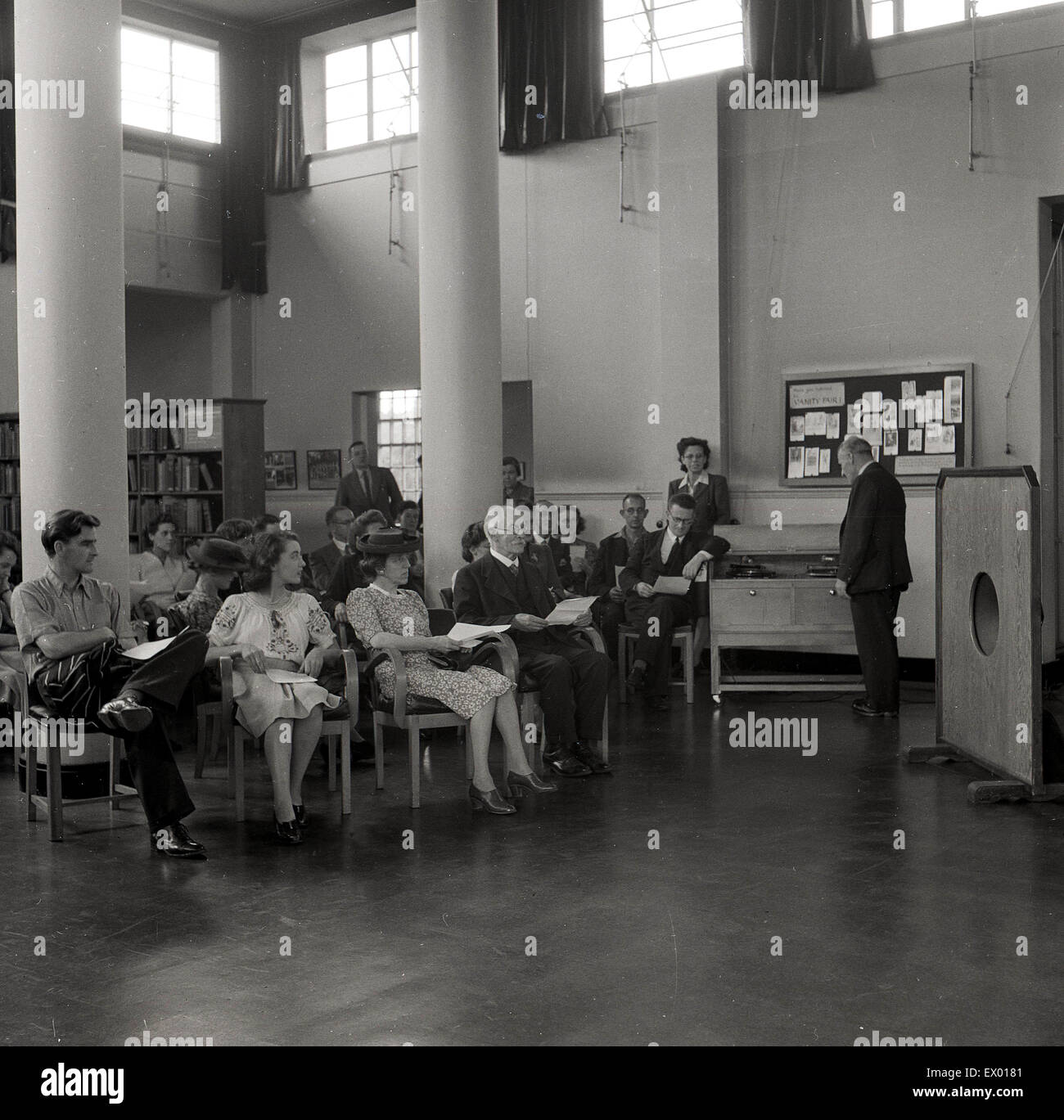 anni '1950, un gruppo di adulti seduti su sedie rigide in un'area a soffitto alto di una biblioteca pubblica che ascolta musica, un grande altoparlante davanti a loro. Un gentiluomo di un record player. Foto Stock