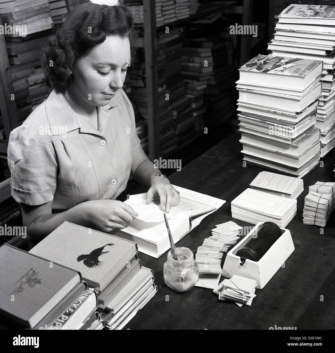 Storico degli anni cinquanta, femmina bibliotecario alla sua scrivania attaccando le operazioni di assunzione e di restituire informazioni foglio all'interno del libro. Foto Stock