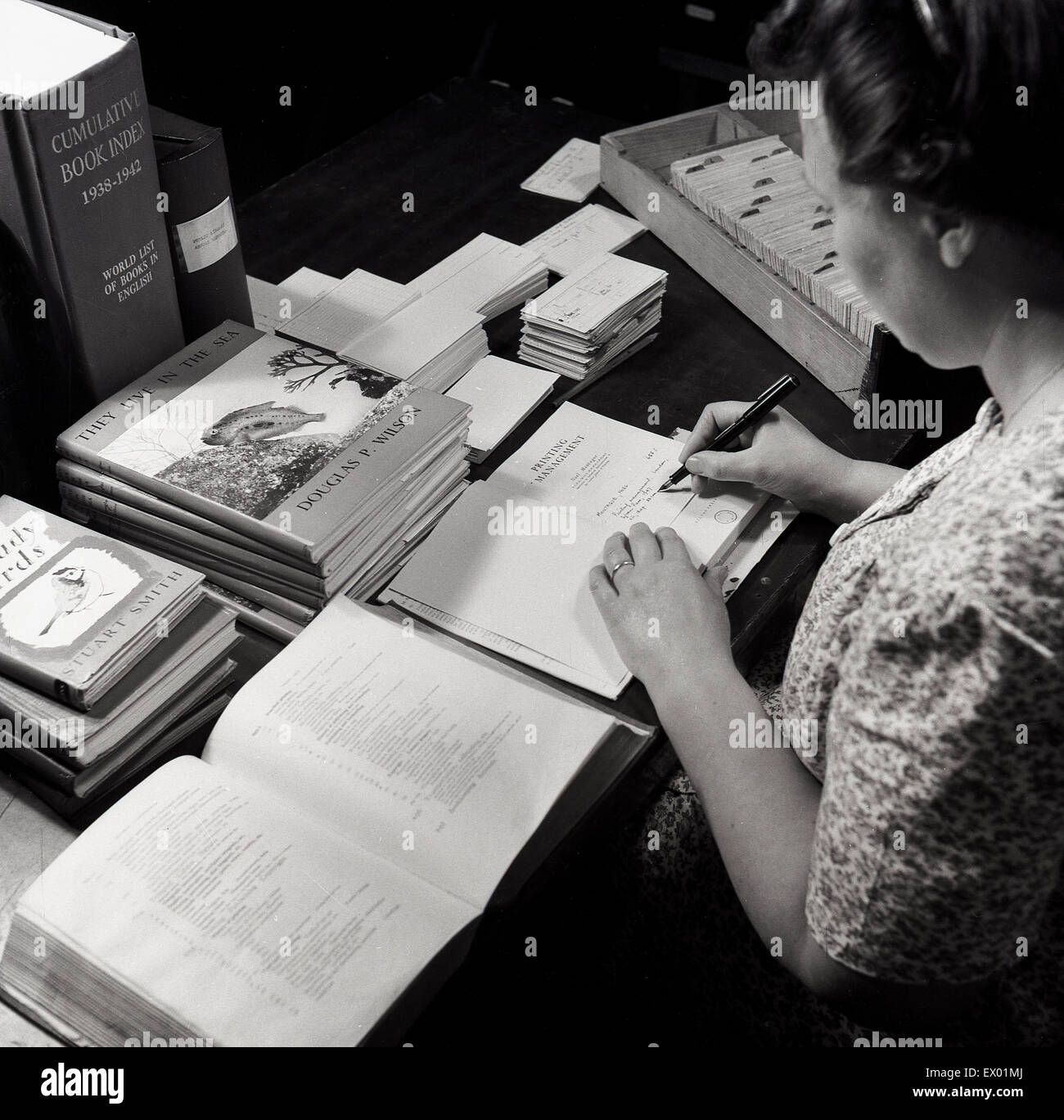 Storico degli anni cinquanta, femmina bibliotecario alla sua scrivania per iscritto le informazioni su una scheda del libro, gli editori e gli autori. Foto Stock