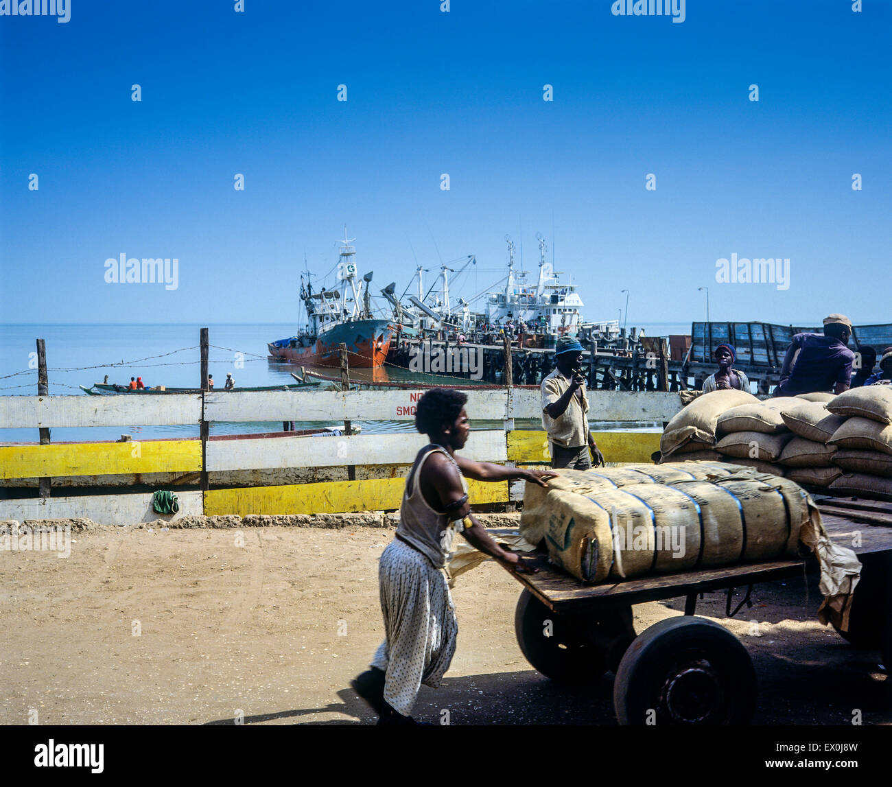 Lavoratori con carretti a sacco, barche da traino ormeggiate al molo in lontananza, porto di Banjul, Gambia, Africa occidentale Foto Stock