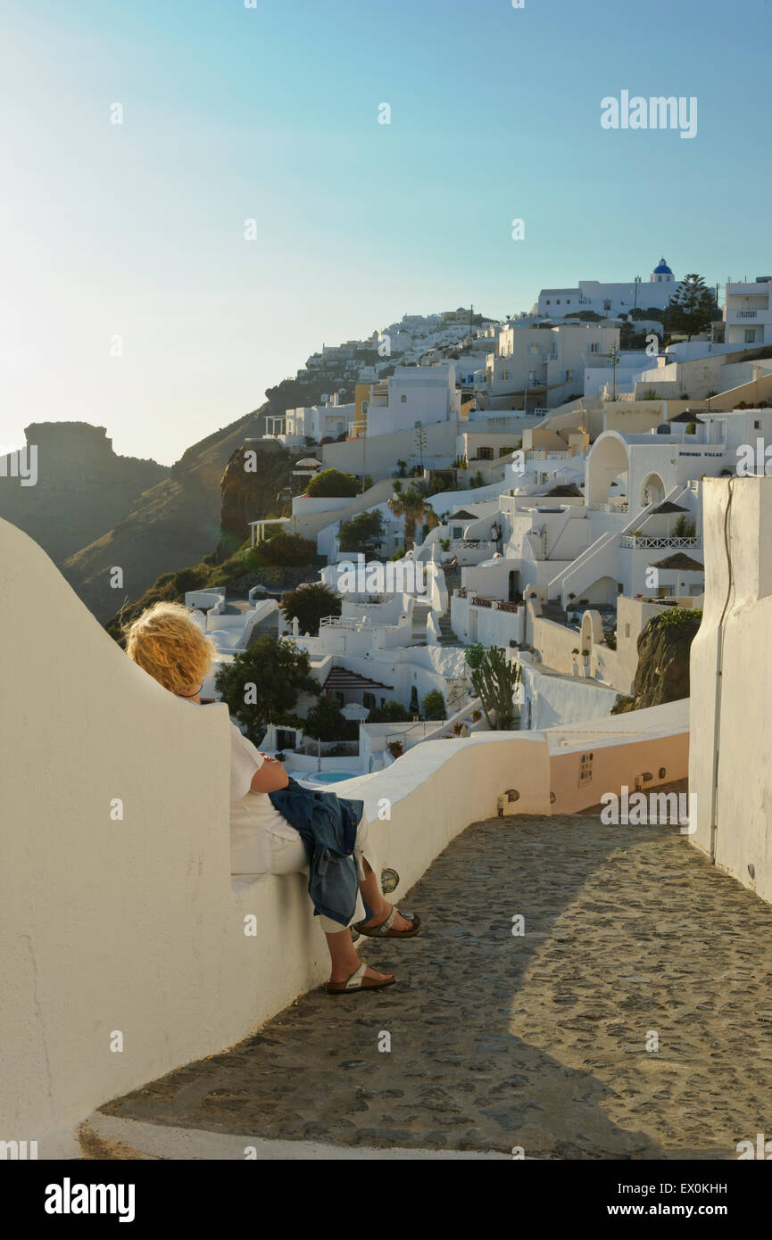 Una donna seduta su una piccola parete ammirando le case sulla Caldera al tramonto a Santorini, Grecia. Foto Stock