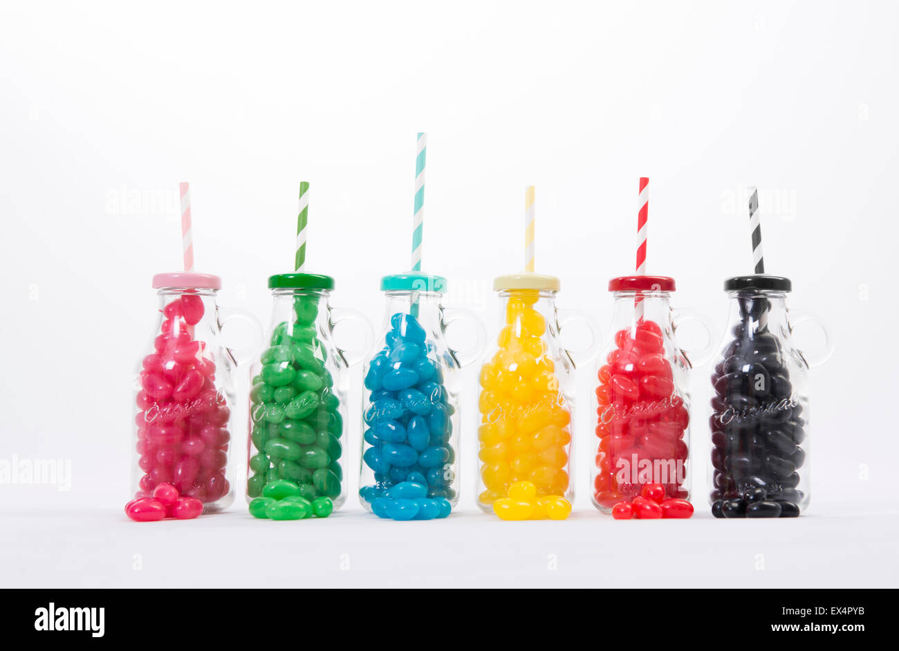 Bottiglie di vetro riempite con differenti jellybeans colorati con corrispondenti coperchi e cannucce, impostare contro uno sfondo bianco. Foto Stock