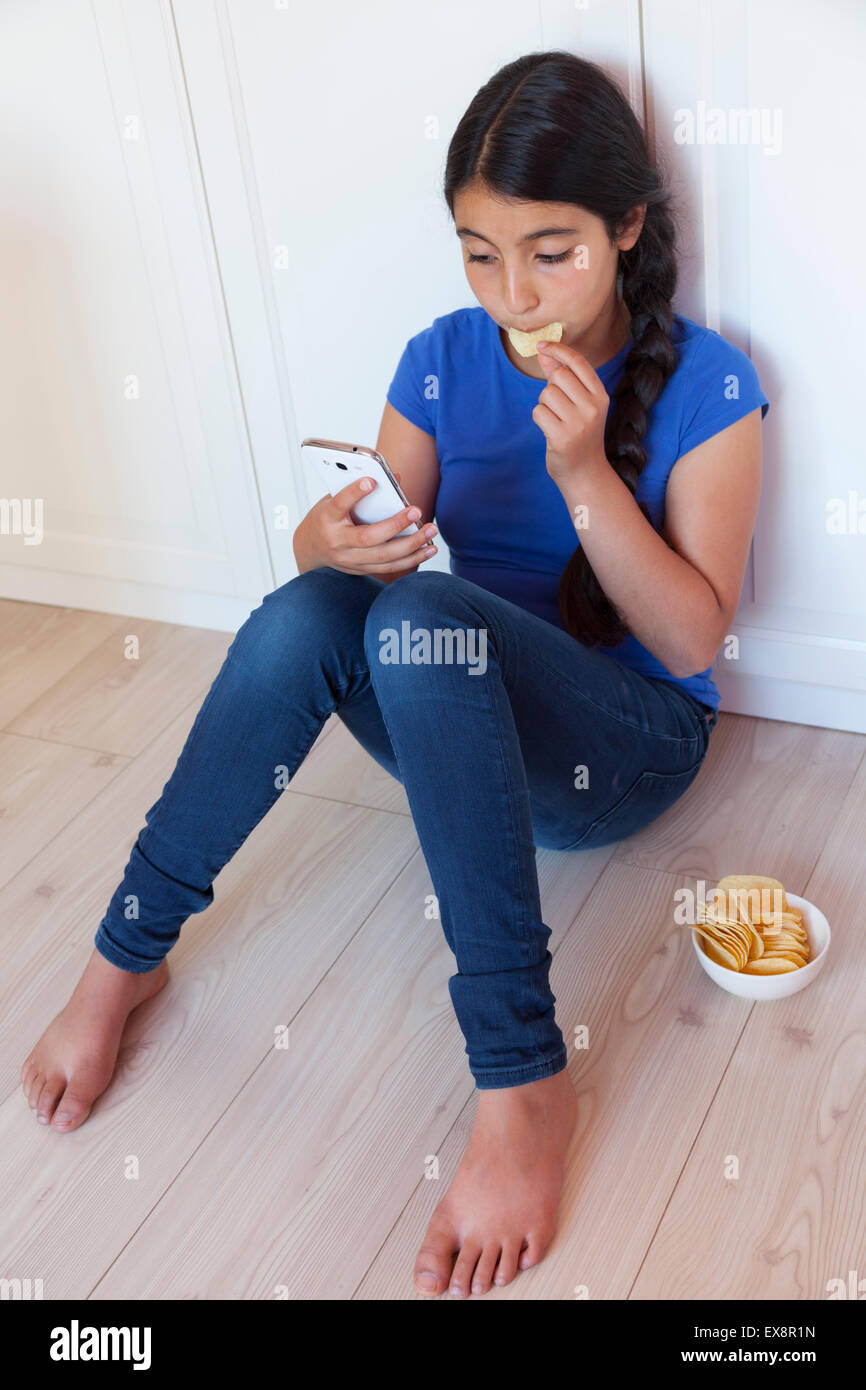 Ragazza adolescente mangiare patatine mentre guardando il suo telefono cellulare Foto Stock