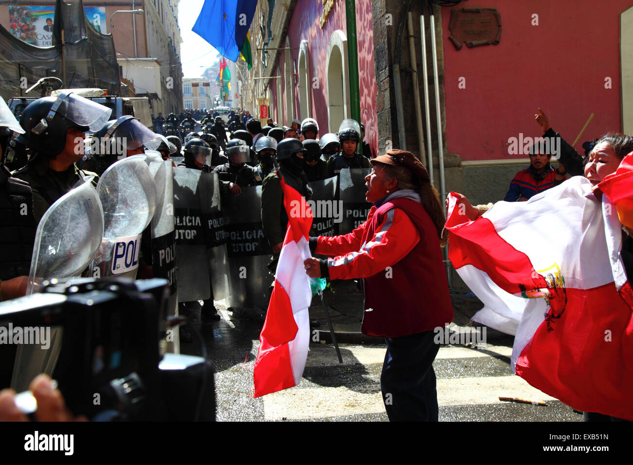 La Paz, Bolivia, 10 luglio 2015. Un manifestante della zona di Potosi si si confronta con la polizia antisommossa durante una protesta del Comitato Civico Potosi e dei suoi sostenitori. Sono a la Paz per chiedere al governo di mantenere le promesse elettorali fatte alla regione in passato. La polizia ha usato gas lacrimogeni per impedire ai manifestanti di entrare in Plaza Murillo (sullo sfondo, dove si trovano il Palazzo Presidenziale e gli edifici del Congresso). Le bandiere rosse e bianche sono le bandiere del dipartimento di Potosí. Credito: James Brunker / Alamy Live News Foto Stock