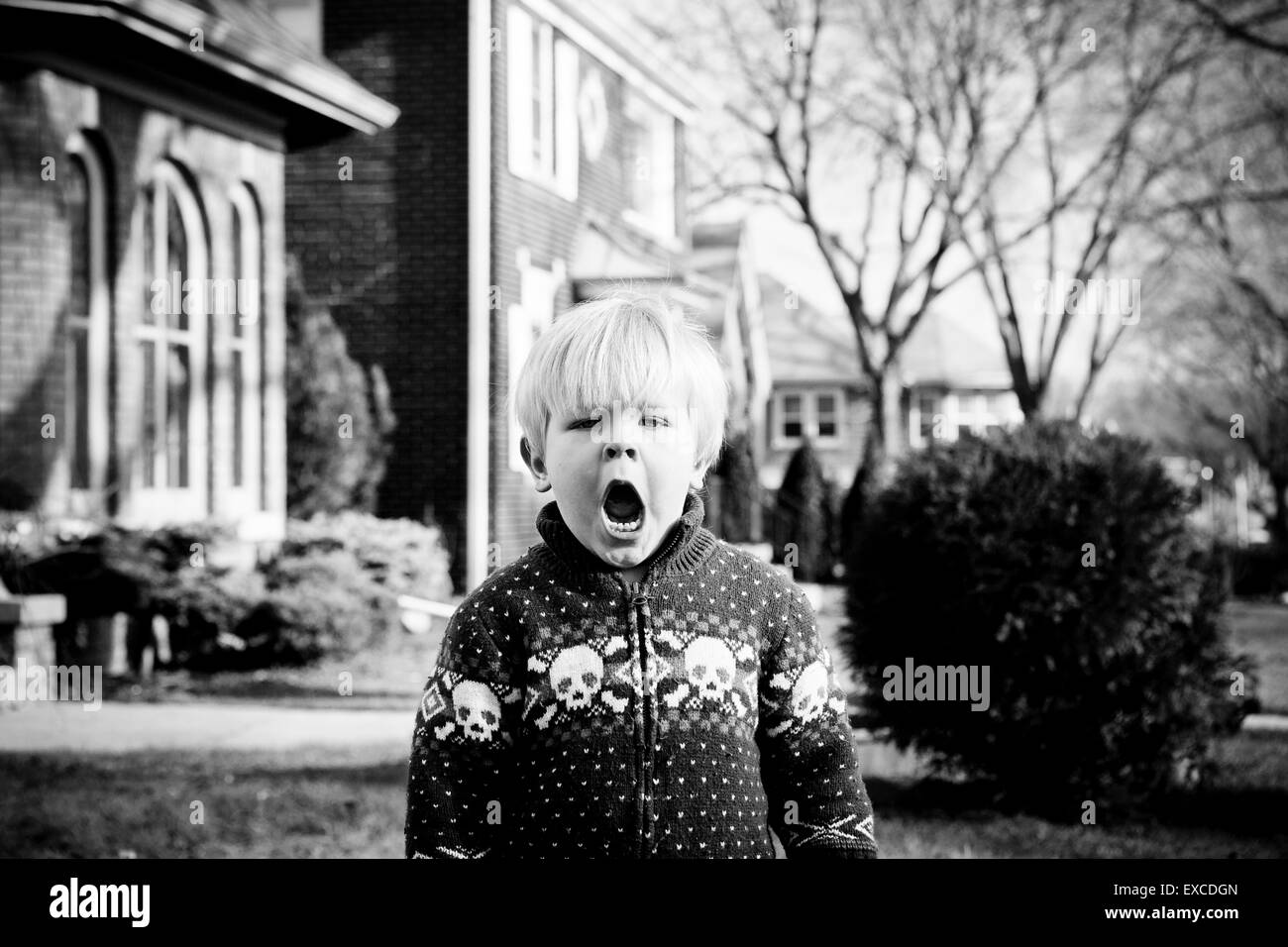 Una scontrosa bionda e giovane ragazzo che indossa un maglione con teschi sorge nel cortile anteriore di una casa in un quartiere. Foto Stock