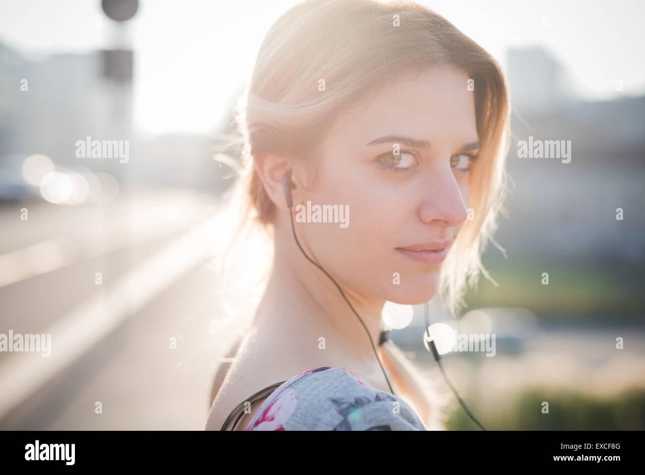 Giovane e bella bionda ragazza caucasica a piedi attraverso la città mentre si ascolta la musica con gli auricolari e uno smartphone, indossando un blu e rosa abito con gonna nera.- tecnologia, musica, serenità e relax concept Foto Stock