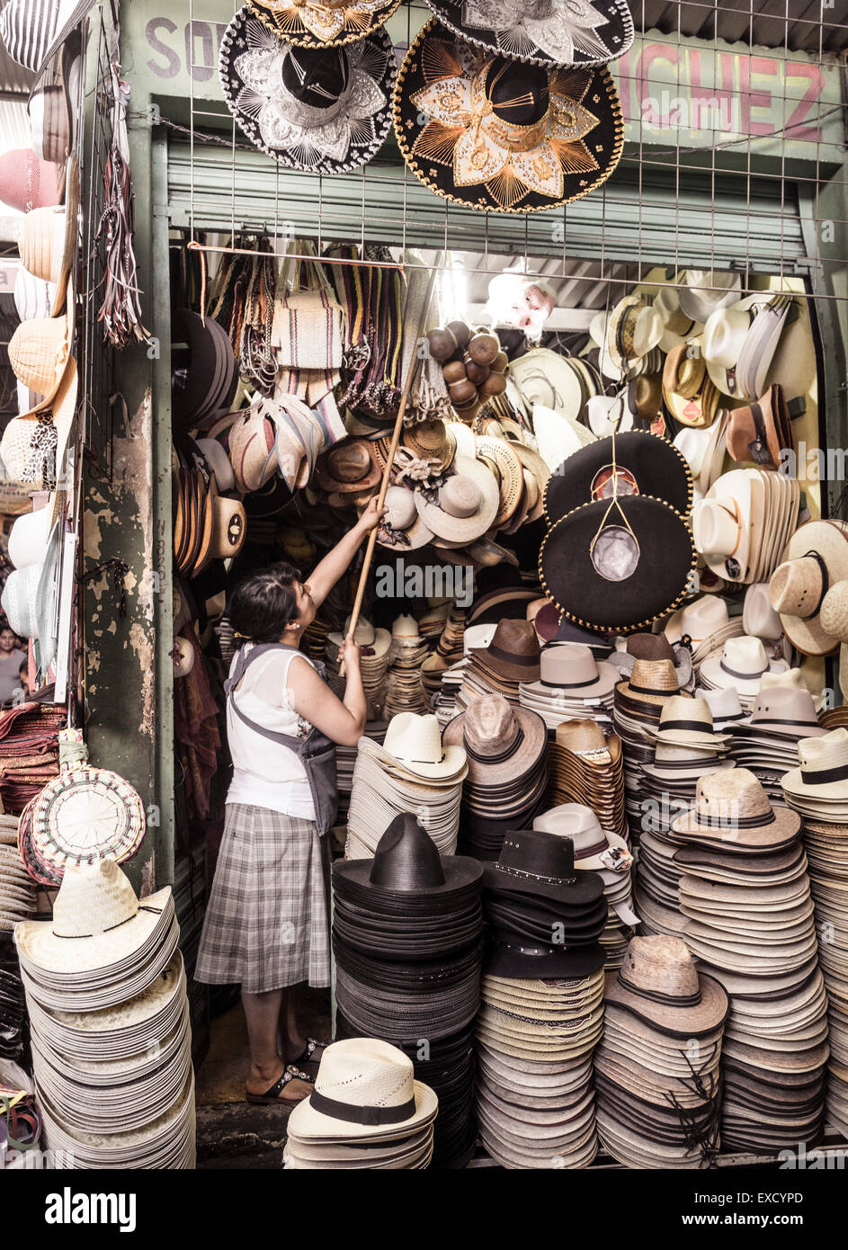 Donna di raggiungere con il polo per appendere sombreros messicani come essa apre la sua piccola hat stallo a un mercato messicano Foto Stock