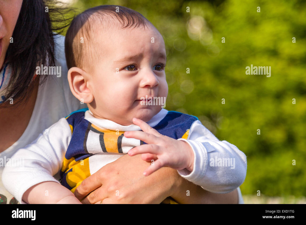 Carino 6 mesi baby con luce marrone capelli in bianco, blu e bruno camicia a maniche lunghe è abbracciato e tenuto da sua madre Foto Stock