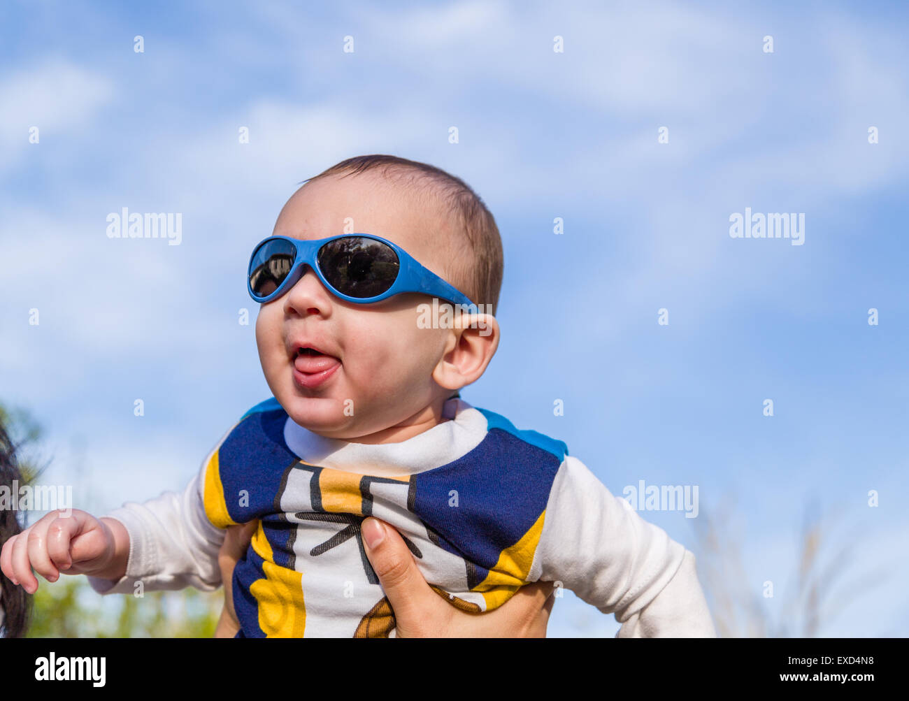 Carino 6 mesi baby con luce marrone capelli in bianco, blu e bruno camicia a maniche lunghe indossando googles blu è abbracciato e tenuto da sua mamma: egli sembra molto felice e sorrisi Foto Stock