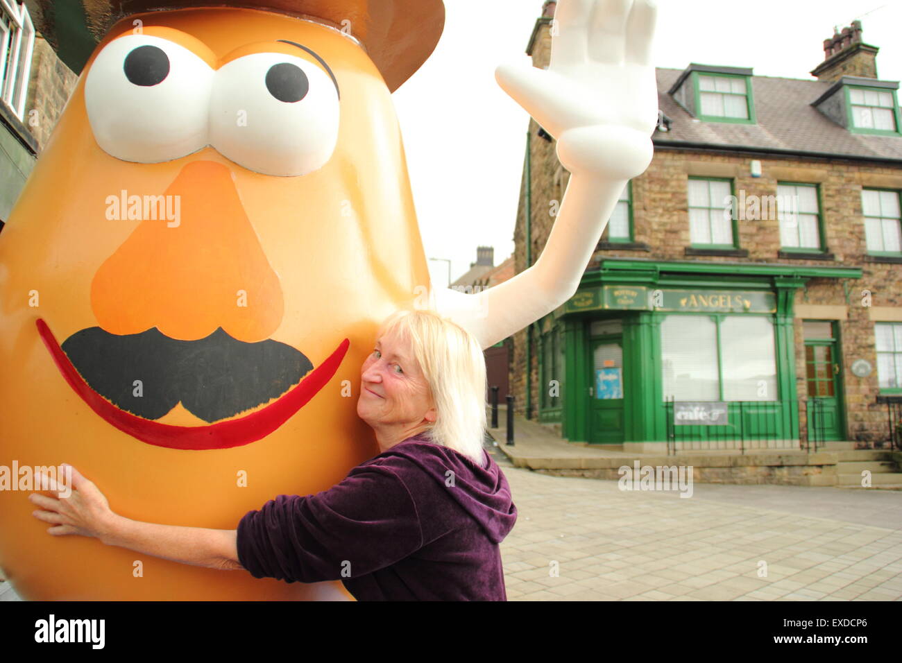Belper, Derbyshire, Regno Unito. 12 Luglio, 2015. Spud ventola, Freda Raphael abbracci un gigante Mr Potato Head che ha ri-apparve in Derbyshire città. La 7ft-alto statua in fibra di vetro è stato regalato a Belper nel 2001 dalla sua città gemelle, Pawtucket, Rhode Island, Stati Uniti d'America. Dopo essere stato ribattezzato "onstrosity" da parte di alcuni locali, il carattere divisivo realizzati titoli nazionali. È stato soggetto ad atti vandalici e bandito. Ora il spud è stata spruced fino da un gruppo locale di giovani e svelato a Belper's Food festival di oggi. Credito: Deborah Vernon/Alamy Live News Foto Stock