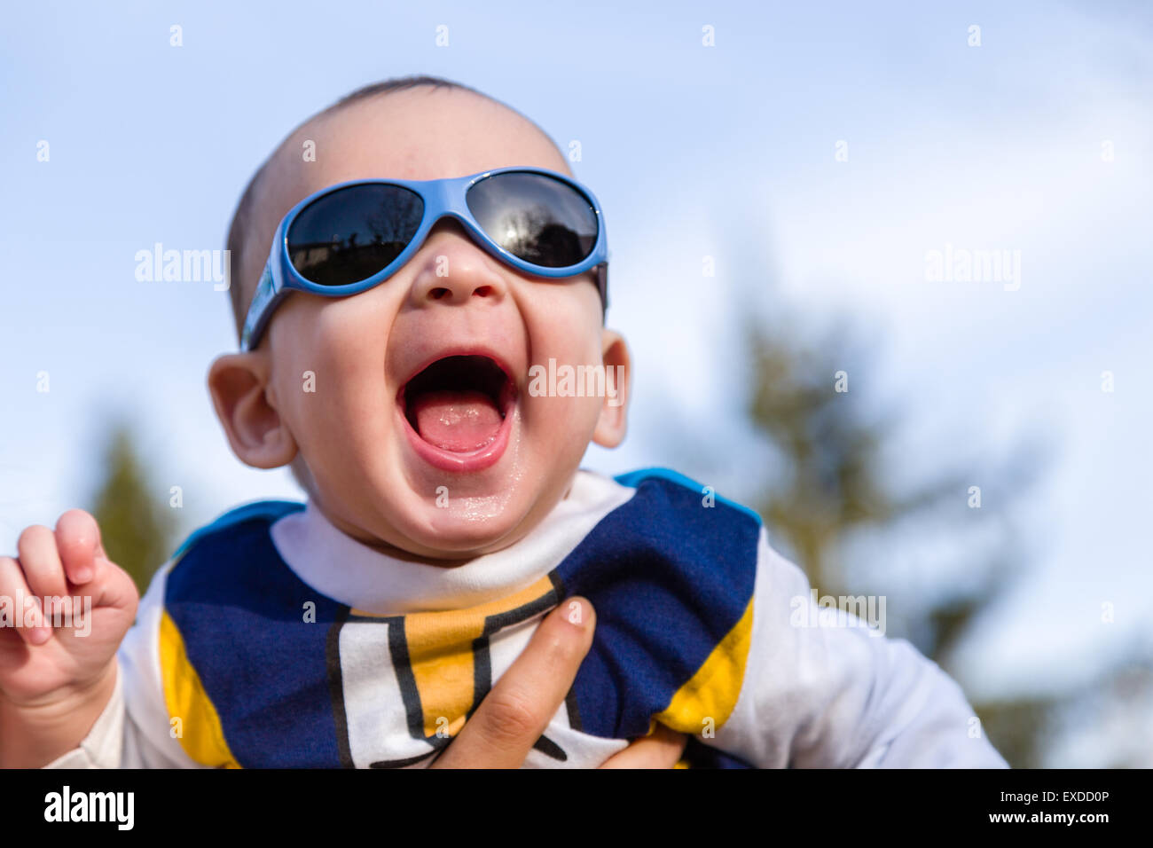 Carino 6 mesi baby con luce marrone capelli in bianco, blu e bruno camicia a maniche lunghe indossando googles blu è abbracciato e tenuto da sua mamma: egli sembra molto felice e sorrisi Foto Stock