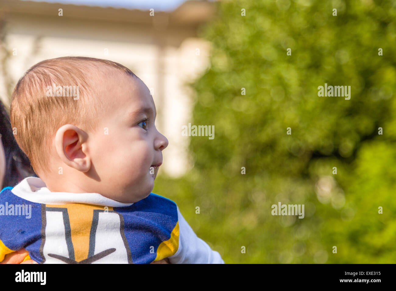 Vista laterale della cute 6 mesi baby con luce marrone capelli in bianco, blu e bruno camicia a maniche lunghe è abbracciato e tenuto da sua madre Foto Stock