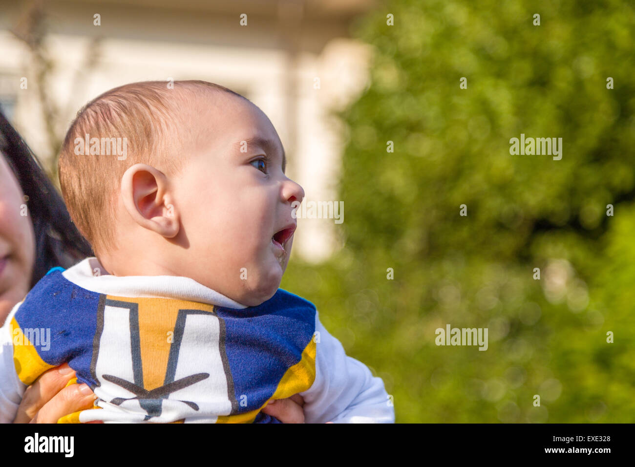 Vista laterale della cute 6 mesi baby con luce marrone capelli in bianco, blu e bruno camicia a maniche lunghe è abbracciato e tenuto da sua madre Foto Stock
