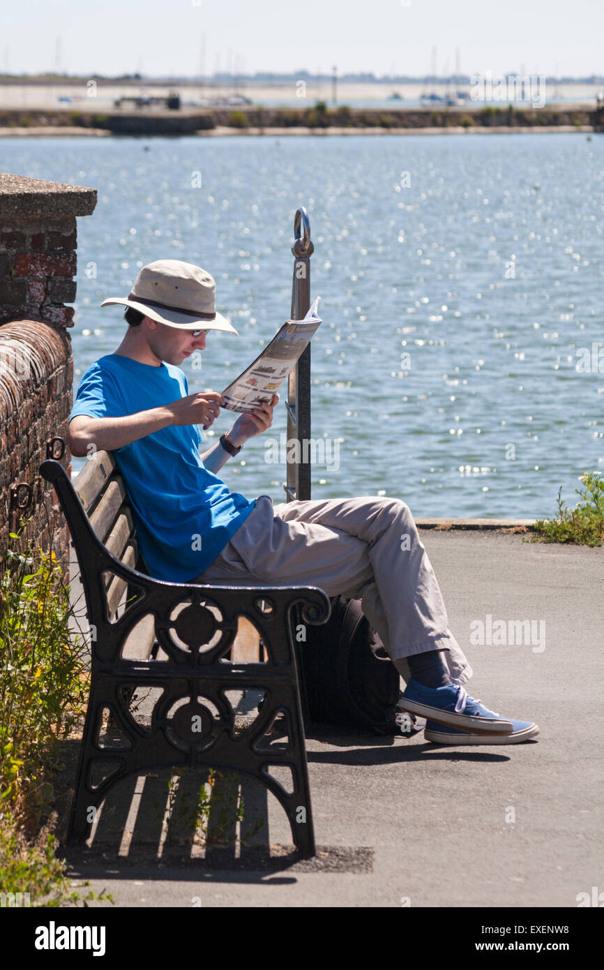 Giovane uomo seduto sul banco la rivista di lettura a Peschici Harbour, Hampshire, Regno Unito in luglio Foto Stock