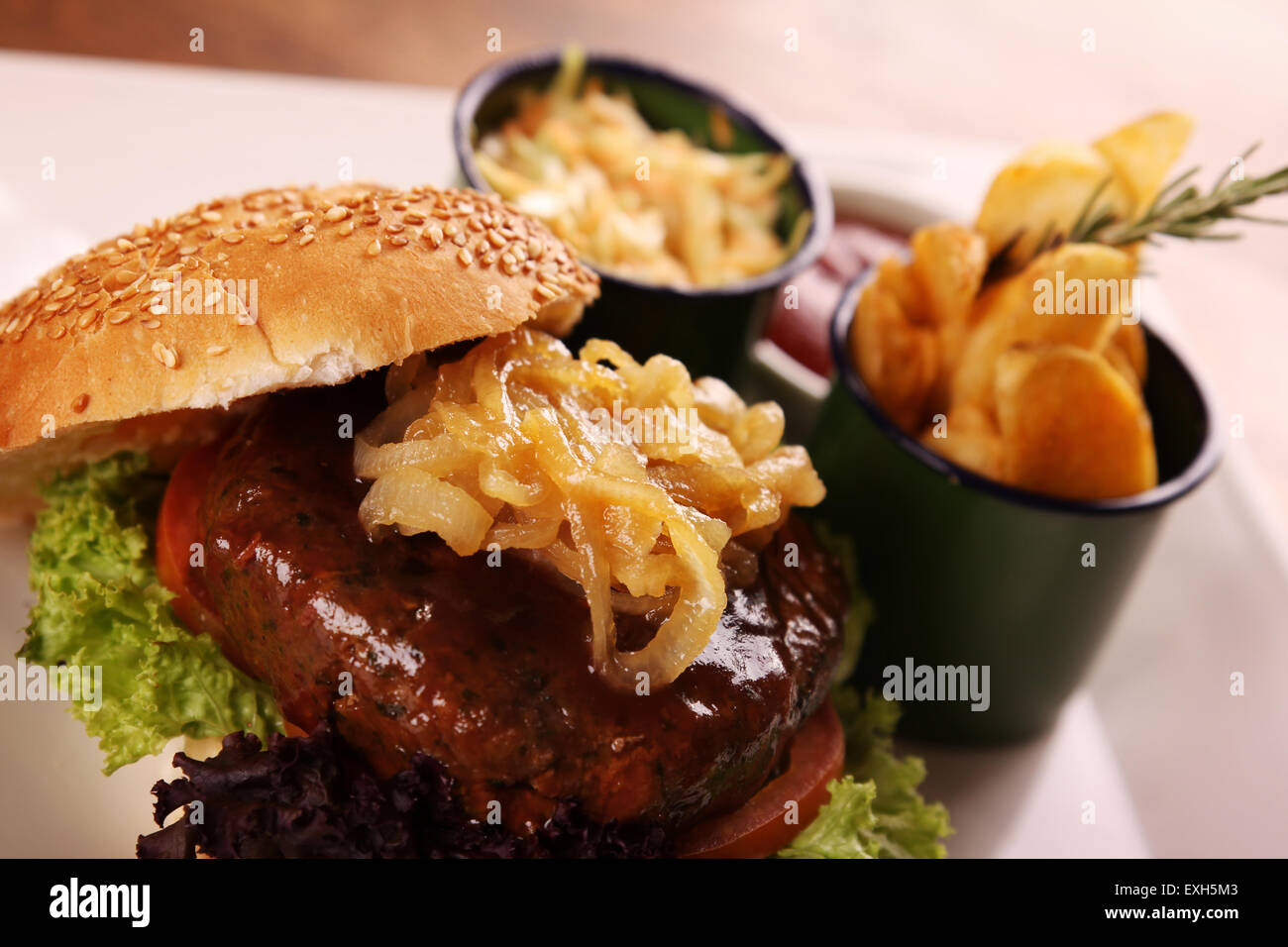 Deliziosa succosa hamburger di manzo con cipolle caramellate e insalata servita con fette di patate su una piastra bianca su di un tavolo di legno. Foto Stock