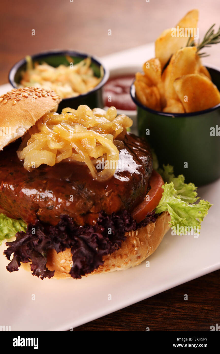 Deliziosa succosa hamburger di manzo con cipolle caramellate e insalata servita con fette di patate su una piastra bianca su di un tavolo di legno. Foto Stock
