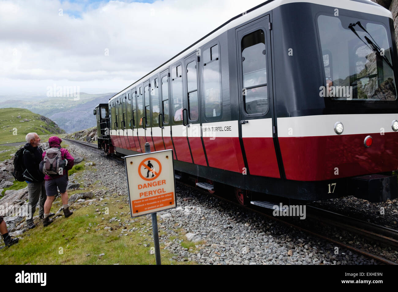 Bilingue segno di pericolo e gli escursionisti in piedi vicino a Snowdon Mountain Railway con il treno che passa sulle vie. Snowdonia Wales UK Foto Stock