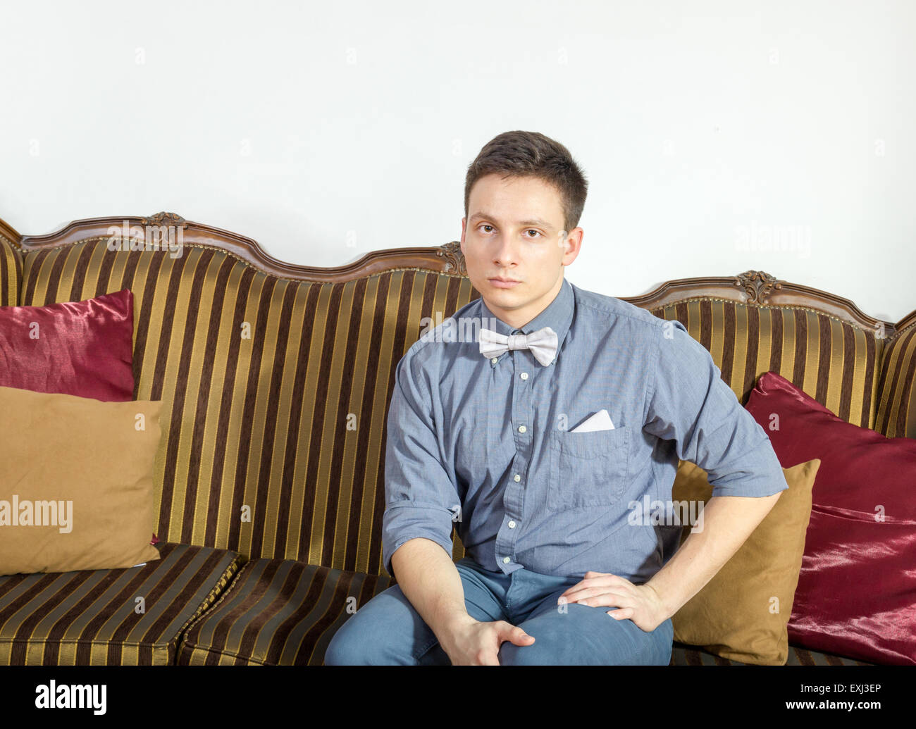 Giovane uomo che pongono seduto su un divano a casa. Modello maschile seduto sul divano di casa. Persona seria nella merda e il filtro bow tie in appoggio su un Foto Stock