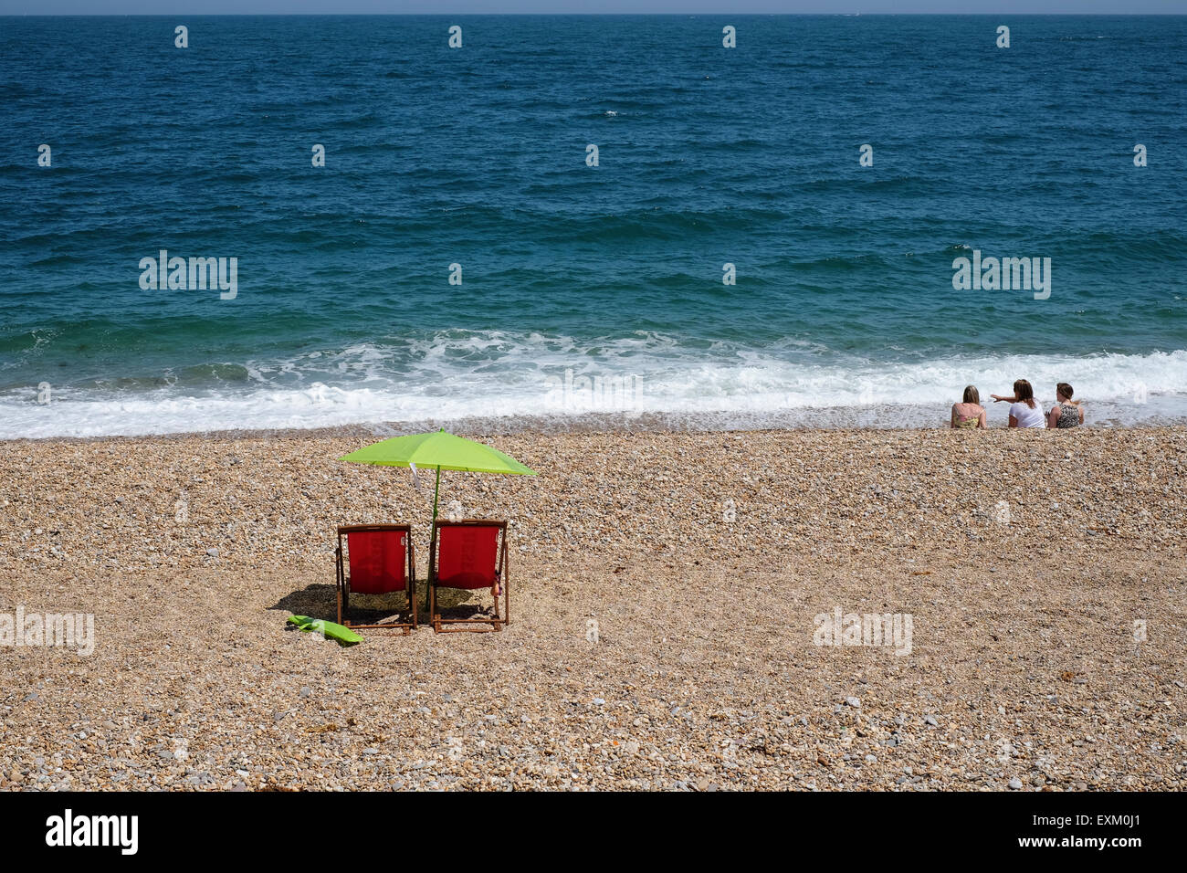 Torcross, Devon, Regno Unito. Rosso di due sedie a sdraio e ombrellone verde sulla spiaggia di ciottoli a Torcross. Foto Stock