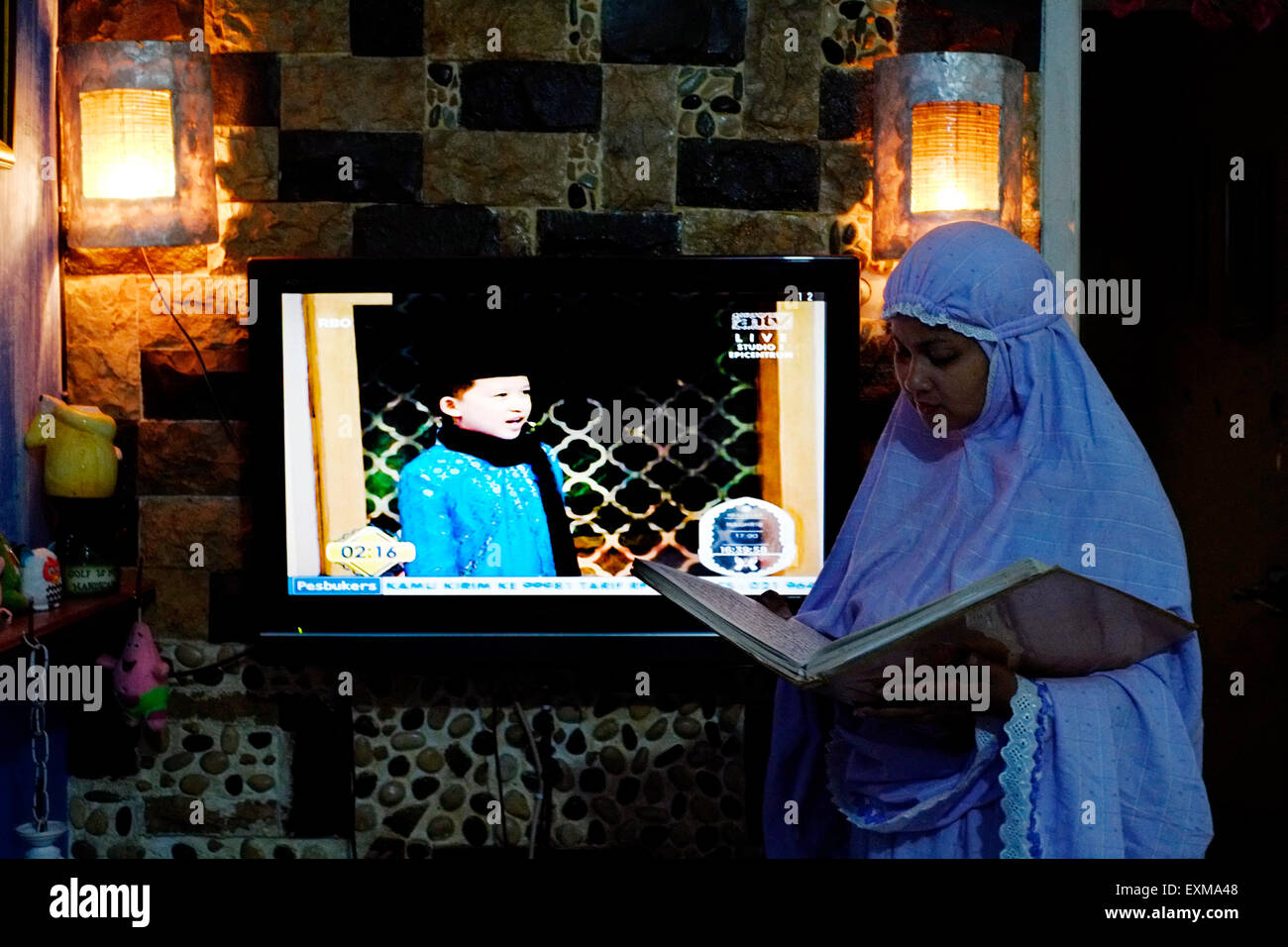 La donna musulmana che indossa il suo abito di preghiera legge il Corano con la televisione di radiodiffusione dietro un programma islamico in Indonesia Foto Stock