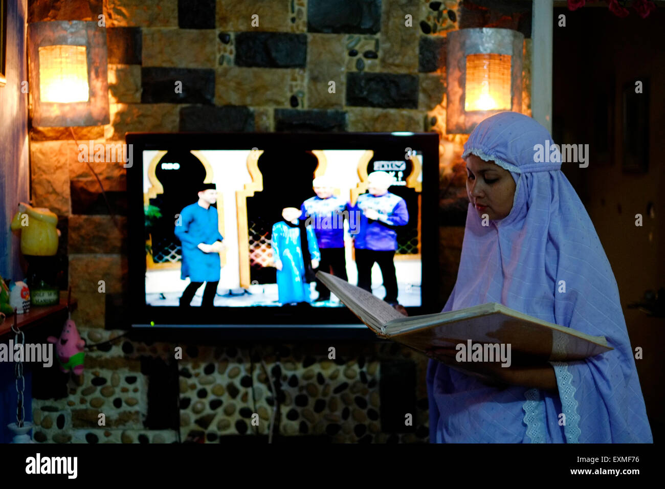La donna musulmana che indossa il suo abito di preghiera legge il Corano con la televisione di radiodiffusione dietro un programma islamico in Indonesia Foto Stock