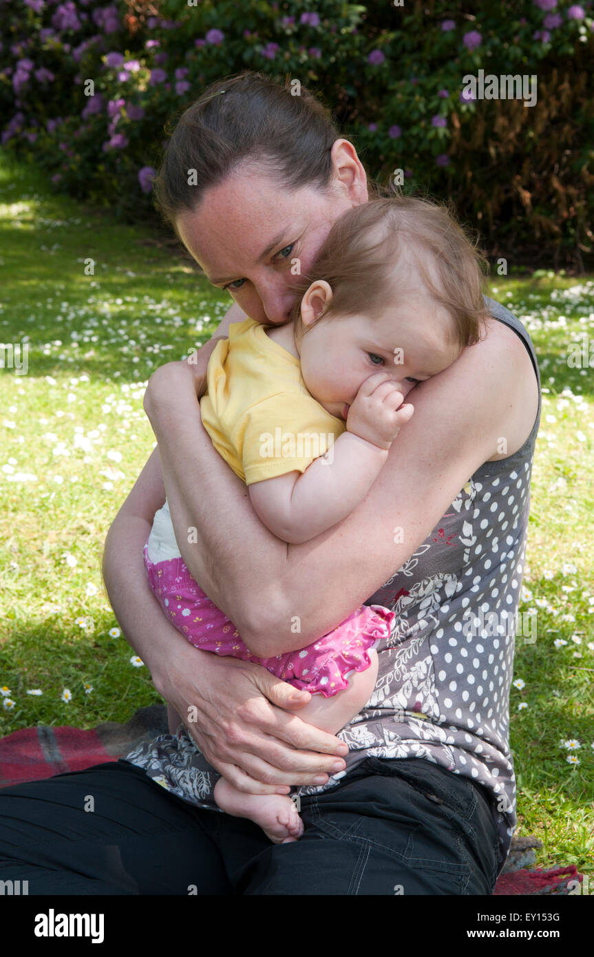 Baby girl succhiare il suo pollice e di essere confortati da sua madre nel giardino Foto Stock