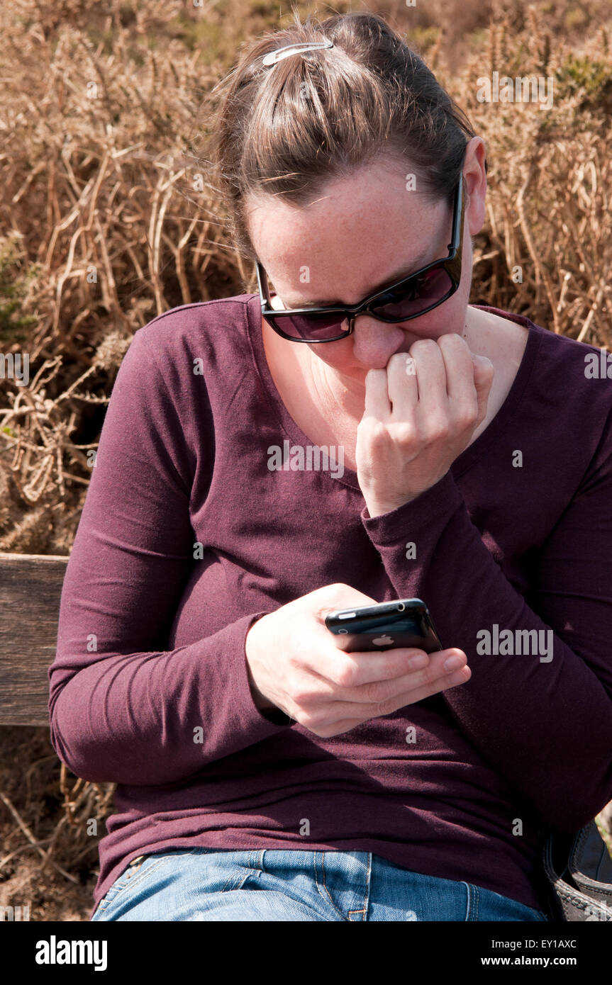 Donna che ha scritto sul suo telefono cellulare guardando preoccupato, seduto su una panca di legno Foto Stock