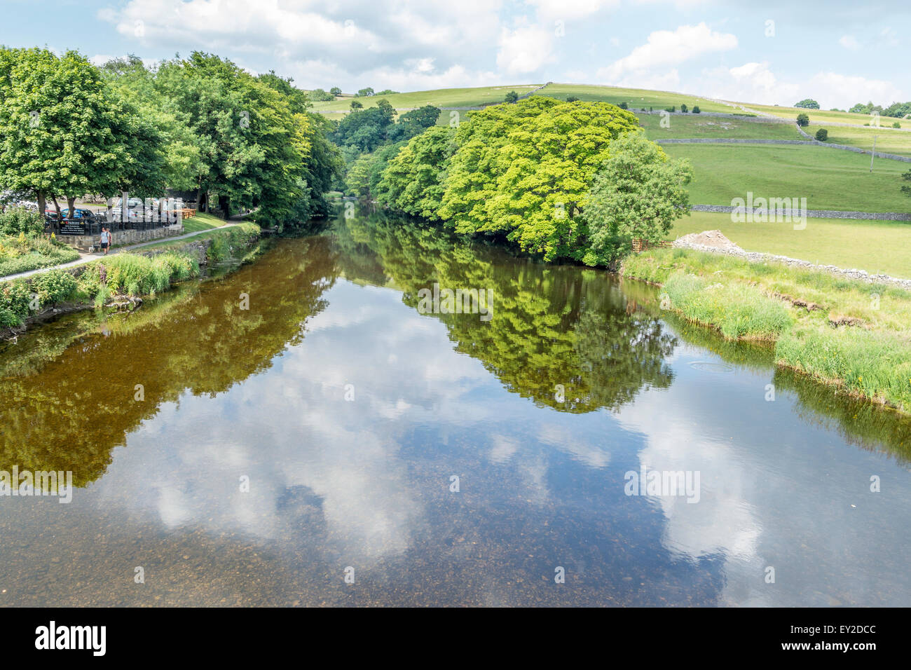Fiume che scorre attraverso una zona boschiva con erba banche su entrambi i lati del fiume, cielo luminoso che è riflessa di ritorno acqua. Foto Stock