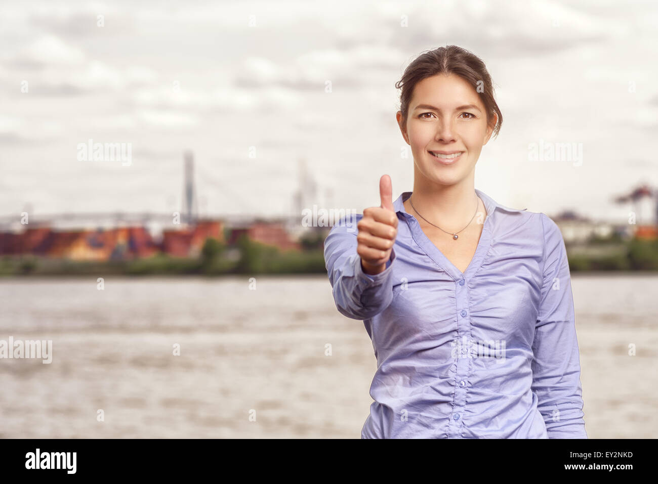 Sorridenti donna attraente dando un pollice in alto gesto di successo e approvazione, corpo superiore ritratto di fronte a un ambiente urbano rive Foto Stock