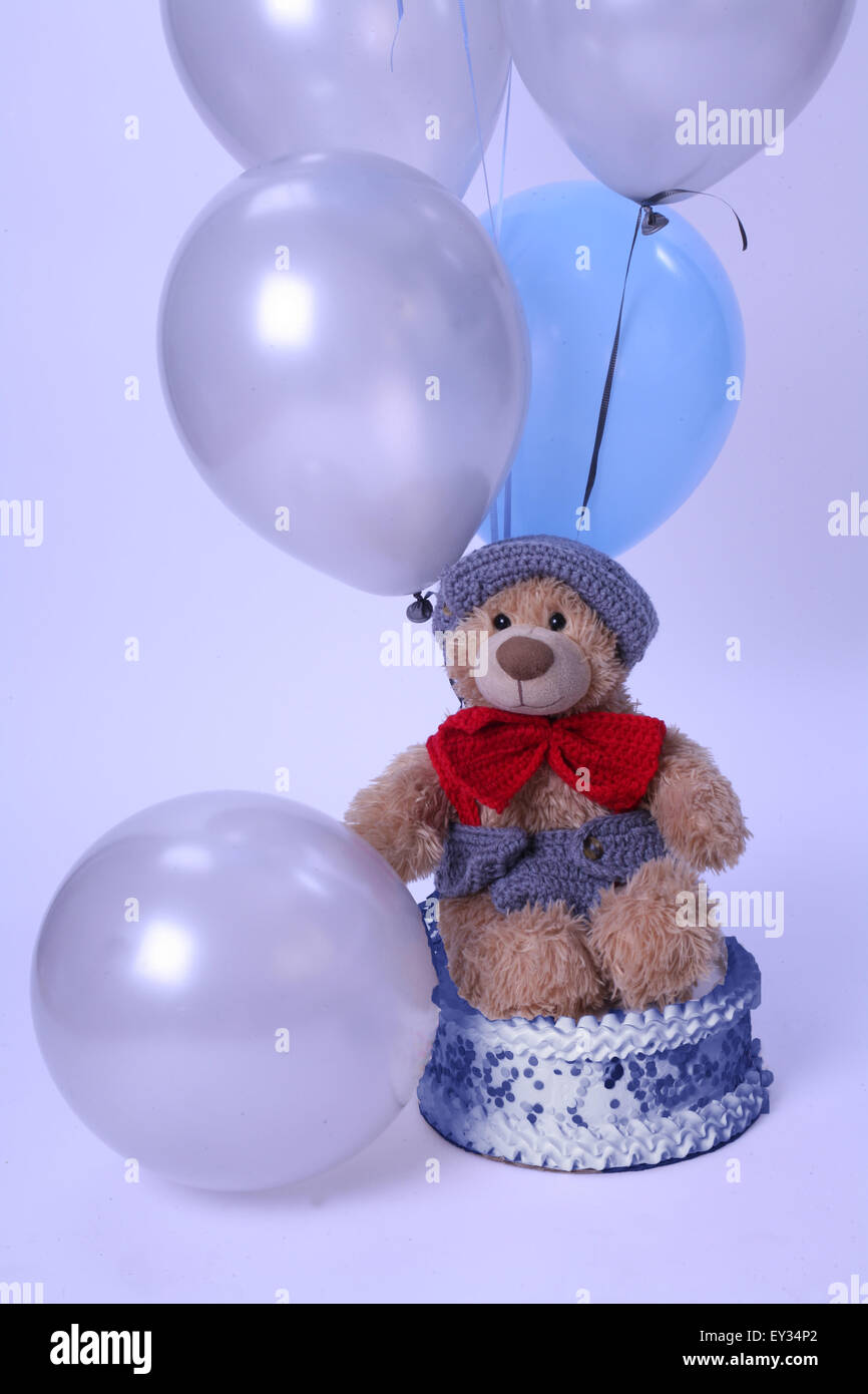 Orsacchiotto di peluche che indossa un bow tie e hat si siede su una torta di compleanno,circondato da palloncini Foto Stock