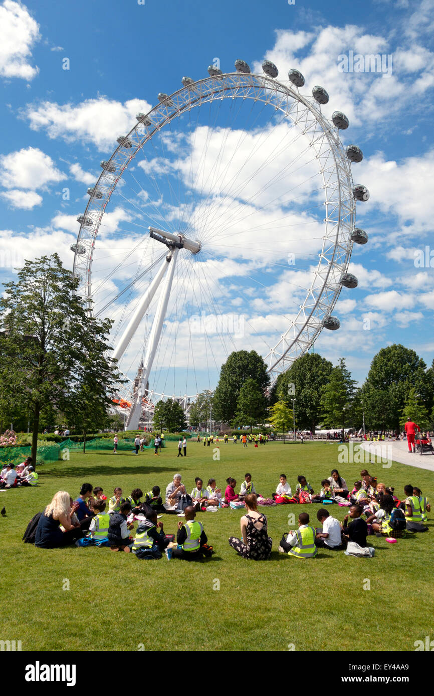 I bambini in gita scolastica sit in Jubilee Gardens sulla South Bank, con l'Occhio di Londra dietro di loro, Londra Inghilterra REGNO UNITO Foto Stock