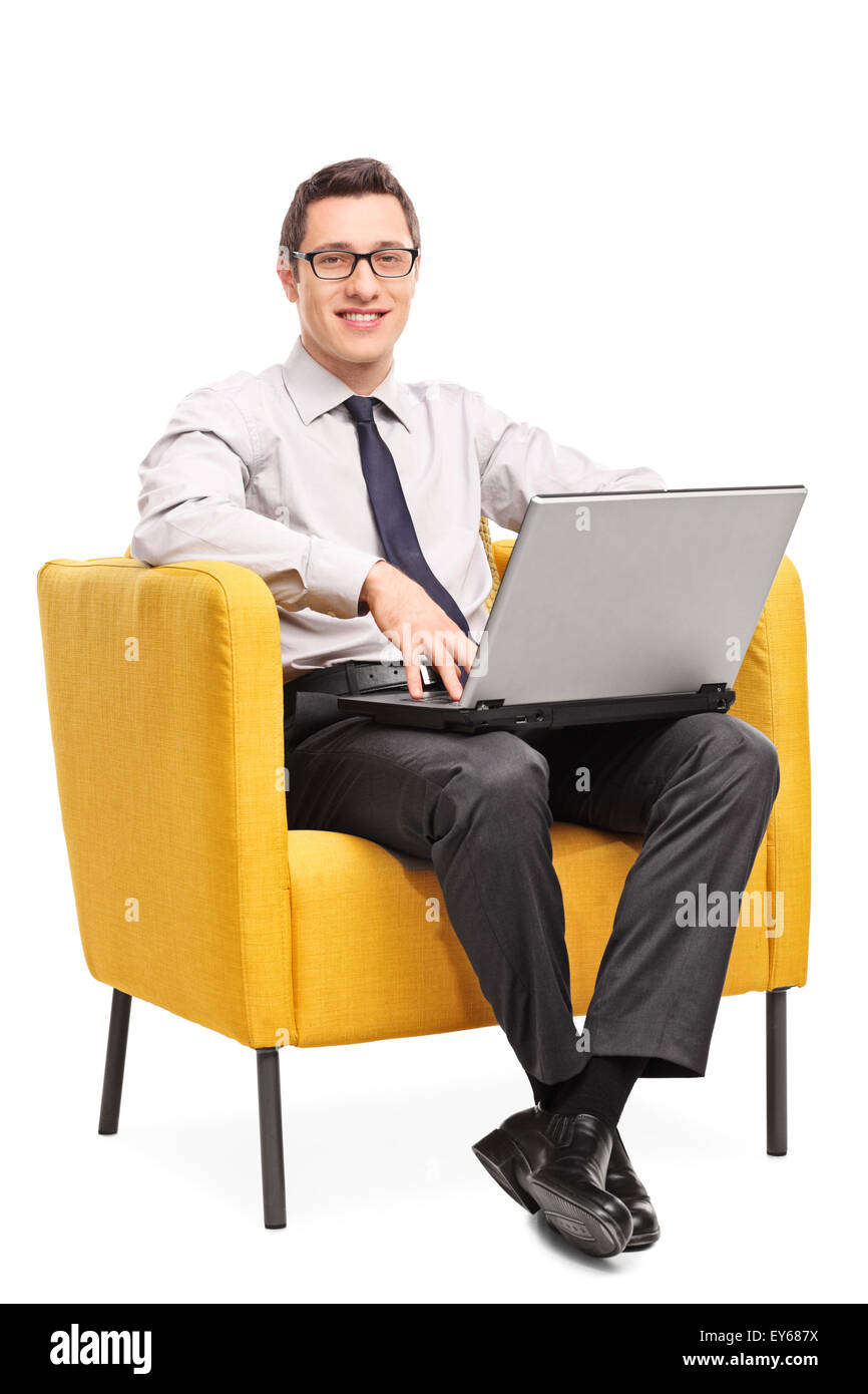 Colpo verticale di un giovane imprenditore allegro lavorando su un computer portatile inserito in una poltrona giallo isolato su sfondo bianco Foto Stock