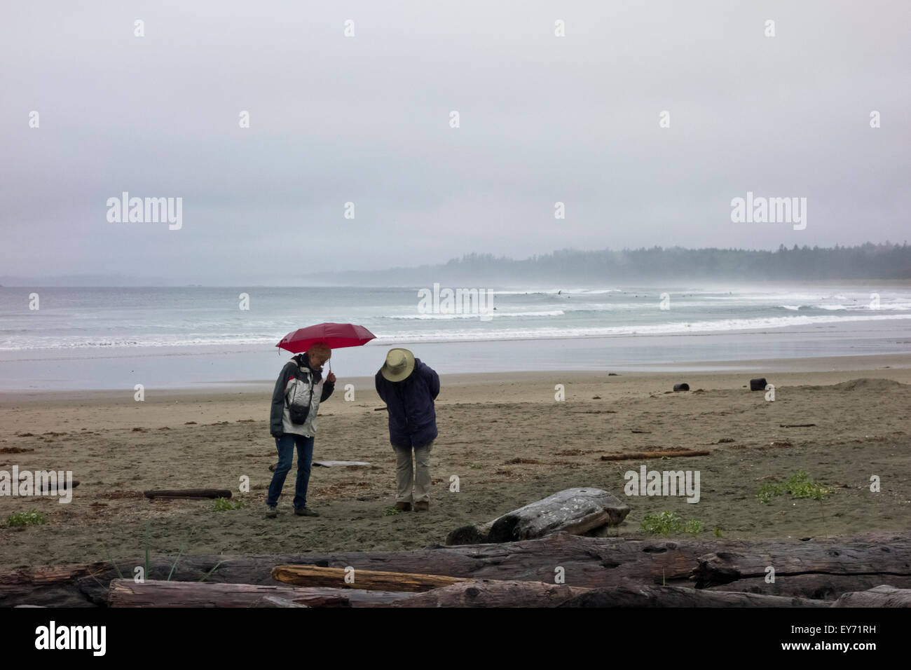 Coppia senior camminando sulla spiaggia Wickanninish in una piovosa giornata d'estate. Surfisti in le onde del mare in lontananza. Long Beach, BC Foto Stock