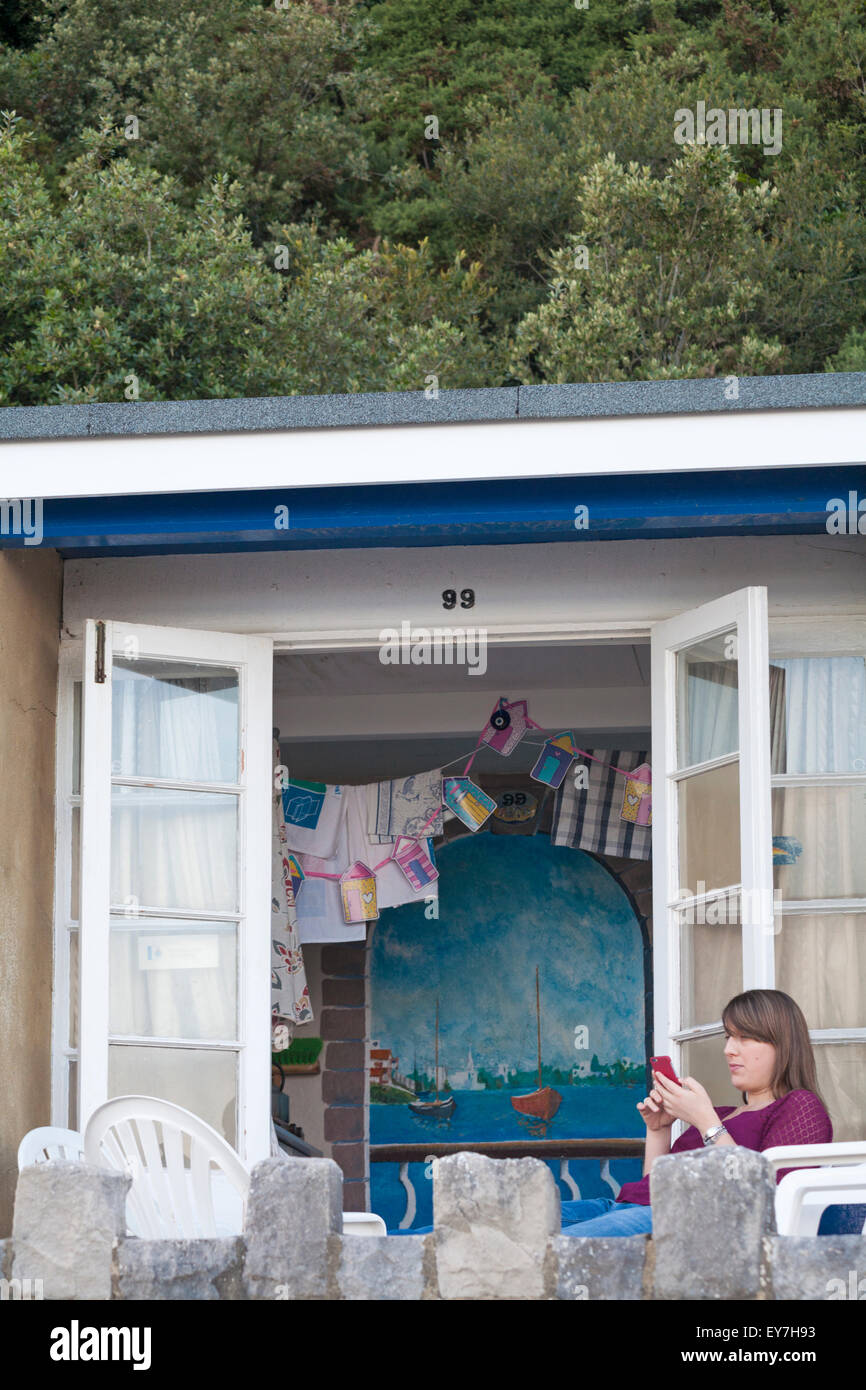 Giovane donna usando il telefono al di fuori seduta colorata beach hut sul lungomare tra cui Canford Cliffs e barene, Poole in luglio Foto Stock