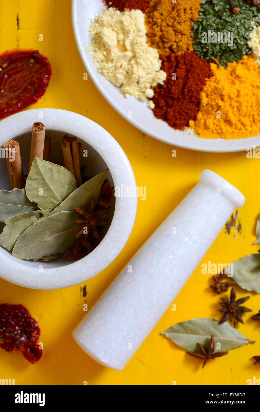 Cucina colorata le spezie e le erbe su un piatto rotondo con bastoncini di cannella, foglie di alloro e anice stellato in un mortaio e pestello su yello Foto Stock