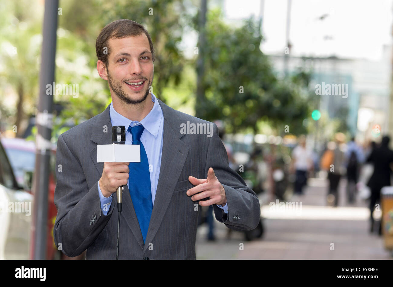 Attraente maschio professionale news reporter indossare tuta grigio tenendo il microfono, parlando con la fotocamera dal contesto urbano Foto Stock